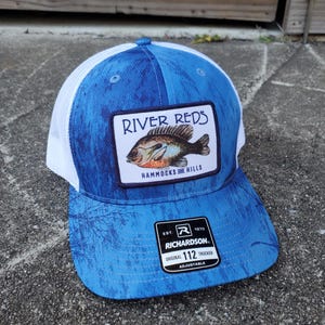 May include: Blue and white trucker hat with a patch featuring a bluegill fish and the text "River Reds Hammocks and Hills". The hat also has a black patch with the text "Richardson 112 Original Trucker Adjustable".