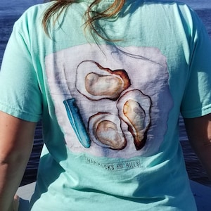 May include: A light blue t-shirt with a white graphic of three oysters and a blue knife. The graphic also includes the text "Hammocks and Hills".