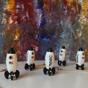 Könnte beinhalten: Fünf Miniatur-Raketen-Ornamente in Weiß und Schwarz mit dem Text "USMC" an der Seite. Die Raketen befinden sich auf einer weißen Oberfläche mit einem verschwommenen Hintergrund aus bunter abstrakter Kunst.