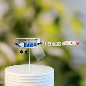 May include: A white wedding cake topper featuring a blue and white airplane with a bride and groom figurine. The airplane is pulling a banner that reads "MR & MRS SMITH" with a red heart at the end.