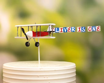 May include: A white and red airplane cake topper with a banner that reads "Oliver is one". The banner is attached to the airplane with a string. The airplane is on a white cake.