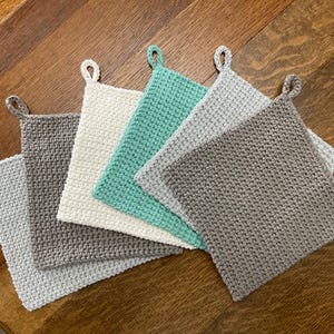 May include: Six handmade crochet potholders in shades of grey, white, and teal. Each square potholder has a loop for hanging. The potholders are made of a textured yarn. The potholders are approximately 20 cm square.