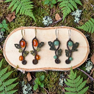 Peut inclure: Deux paires de boucles d'oreilles en macramé composées de cordons marron foncé tressés en forme de feuilles. La première paire comporte des perles orange translucides, tandis que la seconde présente des perles vert clair. Chaque boucle est suspendue à un crochet métallique.