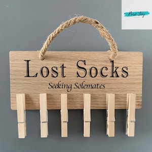 Könnte beinhalten: Ein Holzschild mit dem Text "Lost Socks Seeking Solemates" und einem Seilgriff. Das Schild hat sechs Holzklammern daran befestigt.