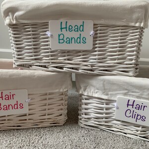 May include: Three white wicker baskets with fabric liners and labels. The top basket is labelled "Head Bands" in teal lettering. The middle basket is labelled "Hair Bands" in pink lettering. The bottom basket is labelled "Hair Clips" in purple lettering.