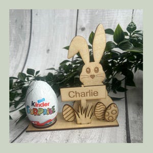 May include: A wooden Easter bunny ornament with the name "Charlie" on a sign. The bunny is holding a sign and has three decorative eggs. A Kinder Surprise egg is next to the bunny.