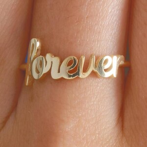 May include: A gold ring featuring the word "forever" in a cursive script. The ring is displayed on a finger, with the word forming the central design. The ring has a polished, reflective surface.