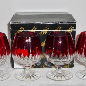 May include: Four red crystal brandy snifters with a cut glass design on the lower portion of the bowl. The glasses sit on a clear, circular base. A black box with gold lettering is behind the glasses.