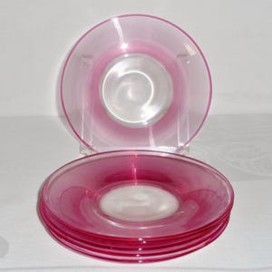 May include: A set of four round glass plates in a vibrant translucent pink hue. Each plate features a smooth, clear center surrounded by a wide rim decorated with fine, concentric circular etched lines that catch the light.