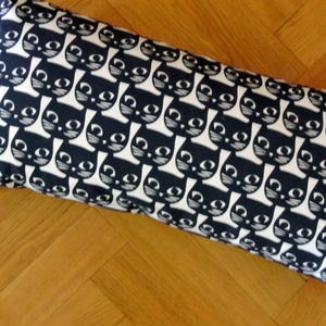 Peut inclure: Un coussin rectangulaire orné d'un motif répétitif de visages de chats noirs stylisés sur fond blanc. Les visages des chats présentent de grands yeux blancs, des pupilles noires, ainsi que des moustaches et des oreilles dessinées au trait.