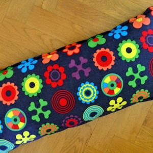 Puede incluir: Un cojín rectangular con una base de tela azul oscuro cubierta por un patrón repetitivo de formas geométricas coloridas, que incluyen flores, círculos y motivos abstractos en forma de cruz en tonos rojo, naranja, amarillo, verde, azul y morado.