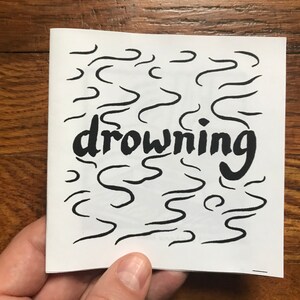 May include: A white booklet with black wavy lines and the word "drowning" written in bold black letters.