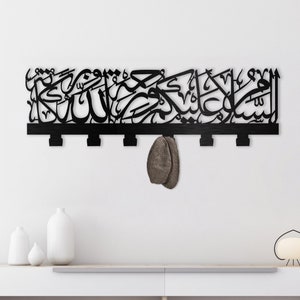 May include: Black metal wall decor featuring Arabic calligraphy and five hooks. The decor is hanging on a white wall above a white shelf with a lamp and vases.