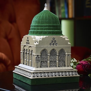 Può includere: Una replica in miniatura di una moschea con una cupola verde e dettagli intricati. Il modello è posto su un tavolo con una superficie scura e un mazzo di rose rosse.