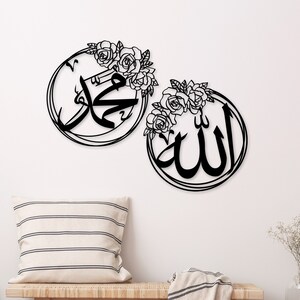K&ouml;nnte beinhalten: Zwei schwarze Wanddekorationen aus Metall mit floralen Akzenten. Die St&uuml;cke zeigen die arabischen W&ouml;rter "Allah" und "Muhammad" in einer dekorativen Schrift.
