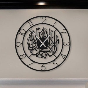 May include: A black metal wall clock with Arabic calligraphy and Roman numerals. The clock is hanging on a white wall above a fireplace with a fire burning inside. The fireplace is in a living room with a grey sofa and a grey armchair.