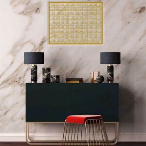 May include: A gold framed wall art with Arabic calligraphy in a modern style. The art is hanging on a white marble wall above a black console table with two black lamps and a red velvet stool.