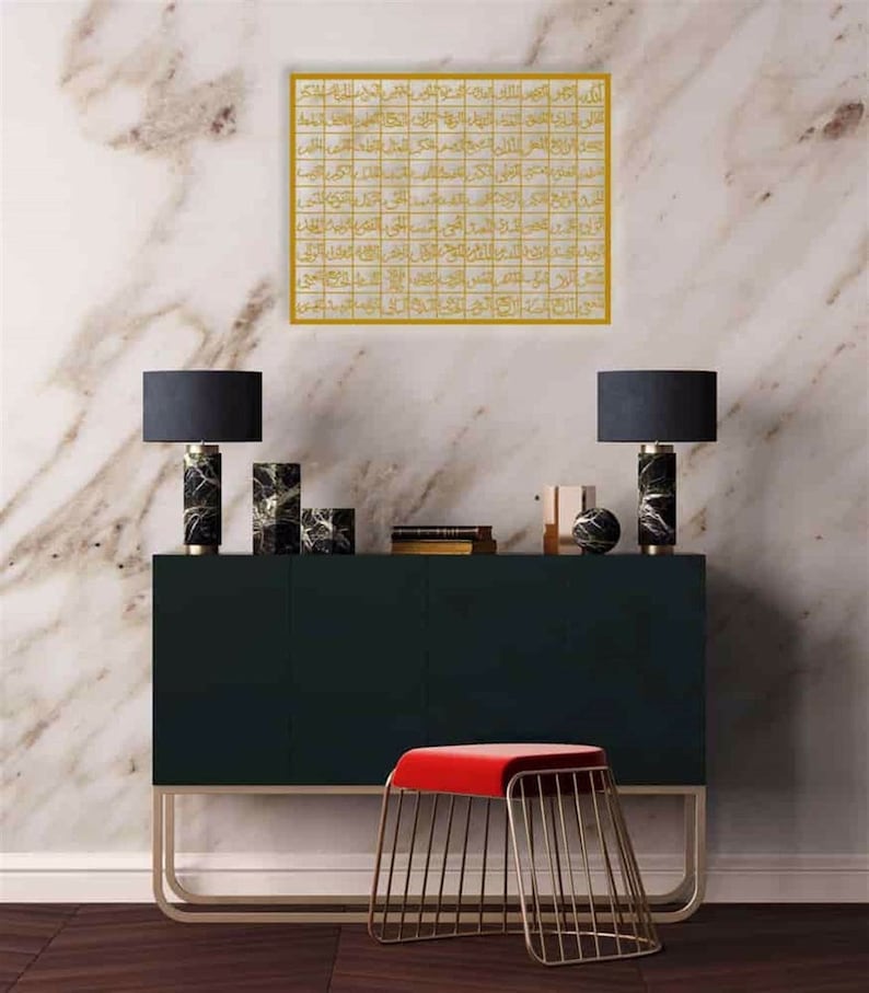 May include: A gold framed wall art with Arabic calligraphy in a modern style. The art is hanging on a white marble wall above a black console table with two black lamps and a red velvet stool.