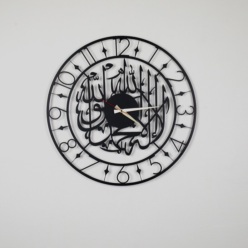 K&ouml;nnte beinhalten: Schwarze, kreisf&ouml;rmige Wanduhr mit arabischer Kalligraphie in der Mitte. Die Uhr hat ein schwarzes Zifferblatt mit wei&szlig;en Ziffern und Zeigern. Das Design ist modern und minimalistisch, geeignet f&uuml;r die Inneneinrichtung.