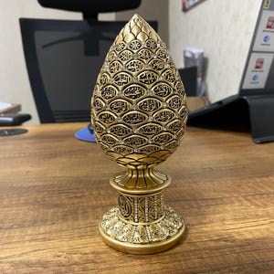 Könnte beinhalten: Ein goldfarbenes, verziertes Dekorationsobjekt in Form eines Tannenzapfens. Die Oberfläche ist mit komplizierten Mustern und Texten bedeckt, mit kleinen, funkelnden Verzierungen. Es steht auf einem detaillierten Sockel, der eine Mischung aus Texturen und Designs zeigt.