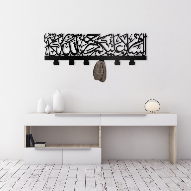 May include: Black metal wall decor featuring Arabic calligraphy and five hooks. The decor is hanging on a white wall above a white shelf with a lamp and vases.