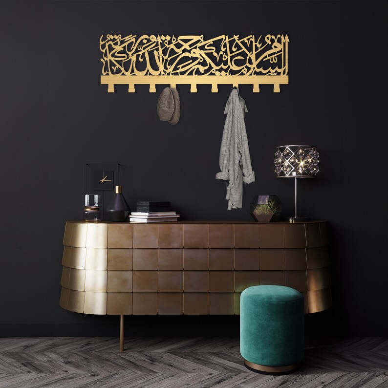 May include: A gold metal wall-mounted coat rack with Arabic calligraphy. The rack has multiple hooks and is hanging on a black wall. A grey scarf and a brown hat are hanging on the hooks. A gold metal console table with a teal velvet stool is in the foreground.