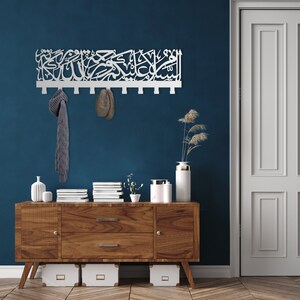 May include: A white wooden wall-mounted coat rack with Arabic calligraphy. The rack has 10 hooks and is mounted on a dark blue wall. A grey scarf and a brown hat are hanging on the hooks. The rack is above a wooden dresser with a white vase and a plant on top.