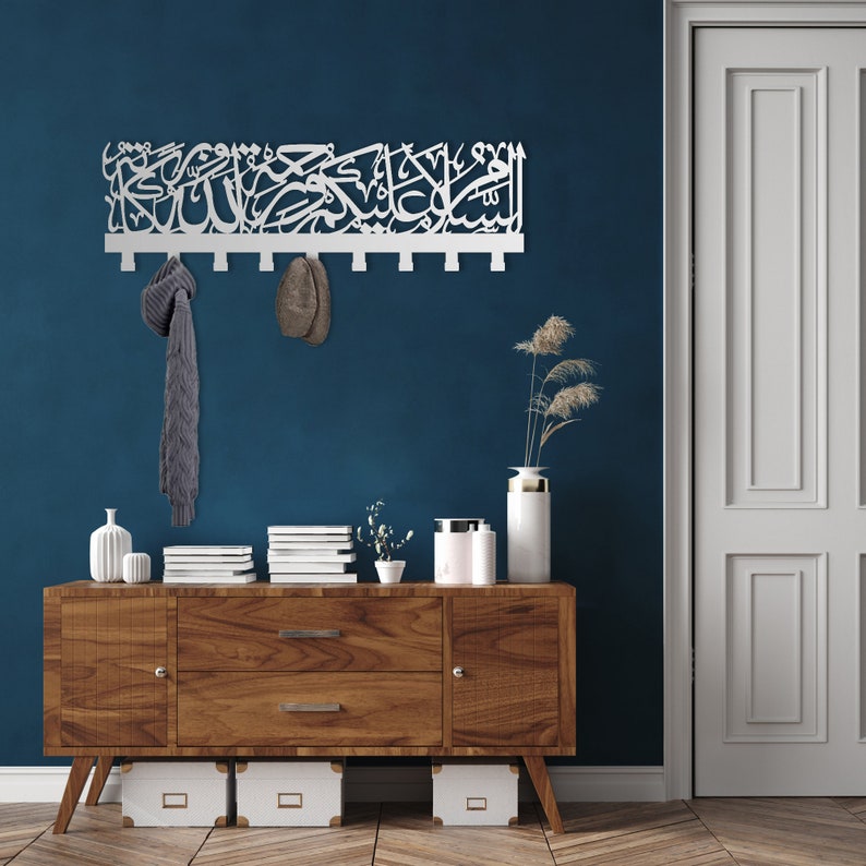 May include: A white wooden wall-mounted coat rack with Arabic calligraphy. The rack has 10 hooks and is mounted on a dark blue wall. A grey scarf and a brown hat are hanging on the hooks. The rack is above a wooden dresser with a white vase and a plant on top.