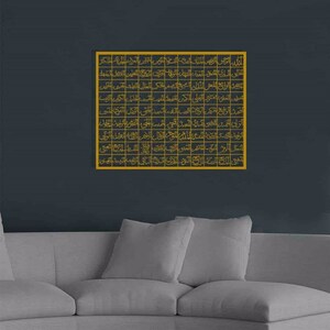 May include: A gold framed wall art piece with Arabic calligraphy in a grid pattern. The art is hanging on a dark gray wall above a gray couch.