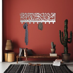 May include: White metal wall-mounted coat rack with Arabic calligraphy. The rack has six hooks and is mounted on a red wall. There is a bench with a brown cushion, a cactus plant, and a lamp in the background.