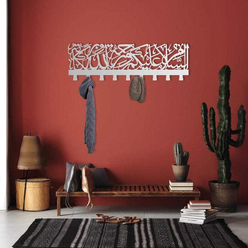 May include: White metal wall-mounted coat rack with Arabic calligraphy. The rack has six hooks and is mounted on a red wall. There is a bench with a brown cushion, a cactus plant, and a lamp in the background.