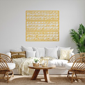 May include: Three gold framed pieces of Islamic calligraphy hang on a white wall above a white couch with pillows and a throw blanket. Two wicker chairs are on either side of the couch. A small wooden table with a plant and a decorative object sits in front of the couch.