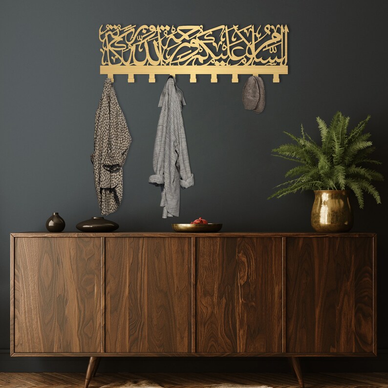 May include: A gold metal wall-mounted coat rack with Arabic calligraphy. The rack has multiple hooks and is hanging on a dark grey wall. There are two items of clothing hanging on the hooks, a grey shirt and a patterned shirt. A brown hat is hanging on one of the hooks. A wooden dresser with drawers is in the foreground. A potted plant is on the right side of the dresser.