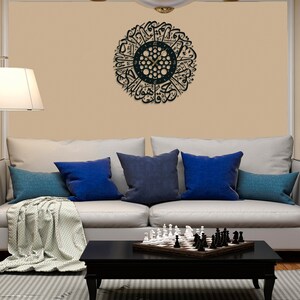 May include: A black wall clock with Arabic calligraphy around the clock face. The clock is hanging on a beige wall above a white couch with blue pillows and a chessboard on a coffee table.