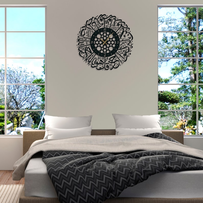 May include: A black and gold wall clock with Arabic calligraphy around the clock face. The clock is hanging on a white wall in a bedroom setting with a bed and windows.