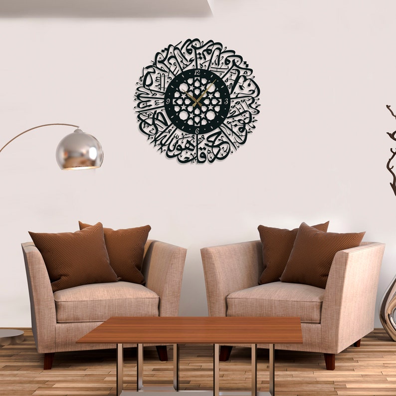 May include: A black wall clock with Arabic calligraphy around the clock face. The clock is hanging on a white wall in a living room setting with two beige armchairs and a wooden coffee table.