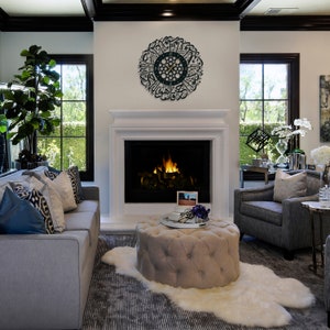 May include: A living room interior with a gray sofa, a gray armchair, a beige ottoman, and a white faux fur rug. A fireplace with a black surround and a roaring fire is in the background. A black clock with Arabic calligraphy is on the wall above the fireplace.