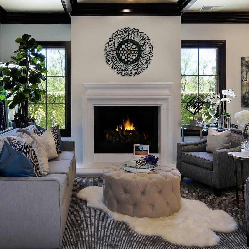 May include: A living room interior with a gray sofa, a gray armchair, a beige ottoman, and a white faux fur rug. A fireplace with a black surround and a roaring fire is in the background. A black clock with Arabic calligraphy is on the wall above the fireplace.