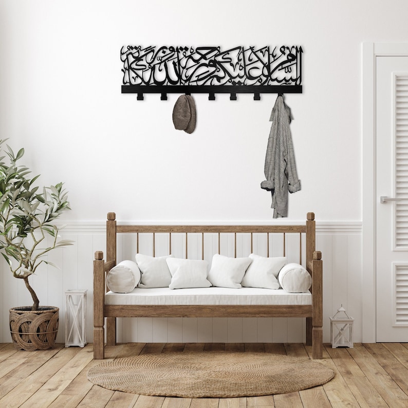 May include: A black metal wall-mounted coat rack with Arabic calligraphy. The rack has six hooks and is hanging on a white wall. A grey jacket is hanging on one of the hooks. A wooden bench with white cushions is in the foreground.