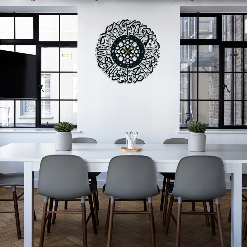 May include: A black and gold clock with Arabic calligraphy around the face. The clock is hanging on a white wall in a dining room with a white table and gray chairs.