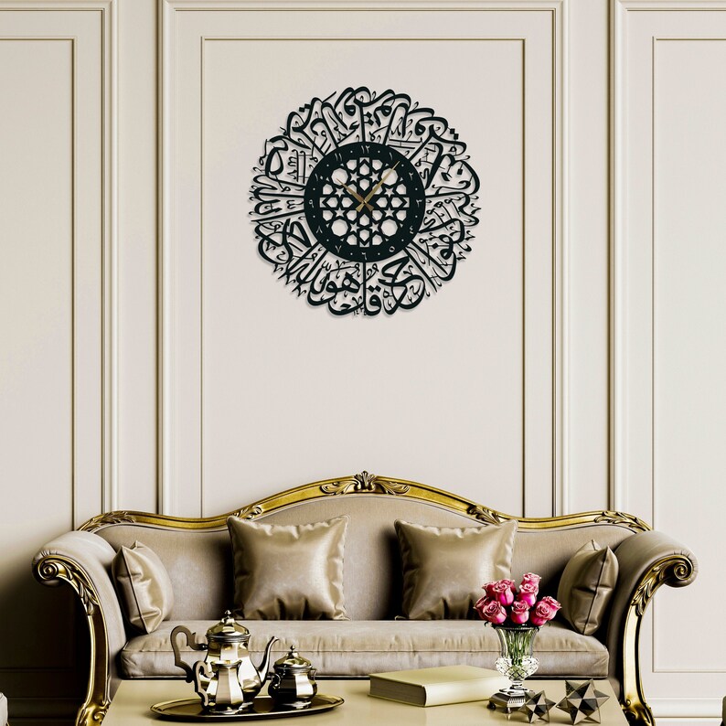 May include: A black metal wall clock with Arabic calligraphy surrounding the clock face. The clock is hanging on a white wall above a beige sofa with gold trim.