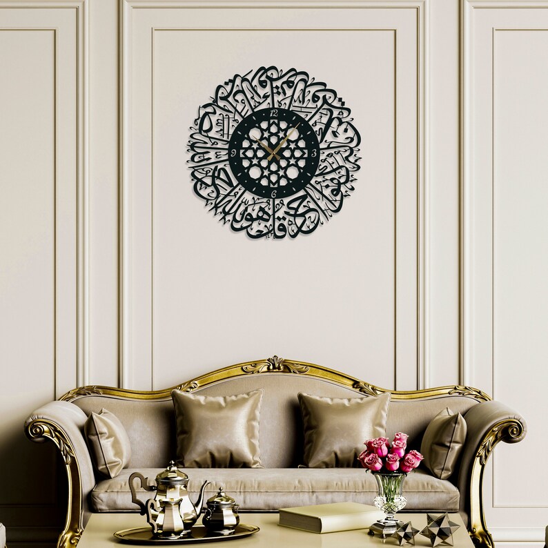 May include: A black wall clock with Arabic calligraphy around the face. The clock has gold hands and a black background. The clock is hanging on a white wall in a living room setting.