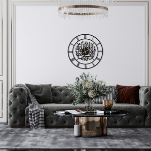 K&ouml;nnte beinhalten: Eine schwarze Metall-Wanduhr mit arabischer Kalligraphie und r&ouml;mischen Ziffern. Die runde Uhr h&auml;ngt an einer wei&szlig;en Wand &uuml;ber einem grauen, getufteten Sofa. Ein Couchtisch und dekorative Gegenst&auml;nde befinden sich davor.