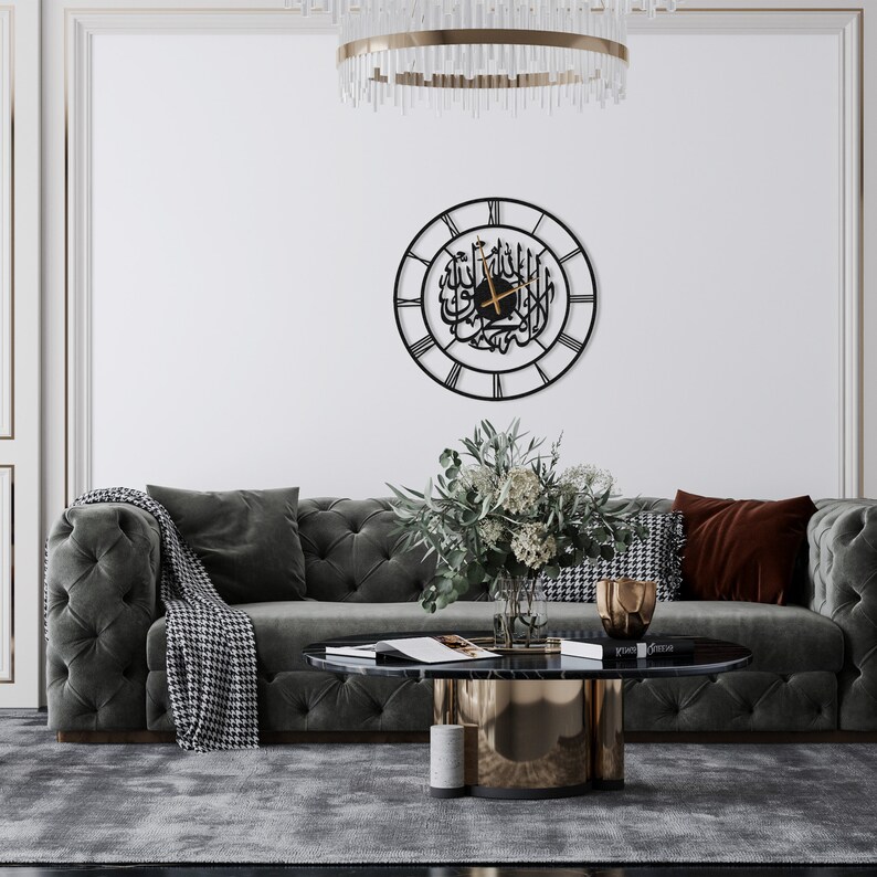 K&ouml;nnte beinhalten: Eine schwarze Metall-Wanduhr mit arabischer Kalligraphie und r&ouml;mischen Ziffern. Die runde Uhr h&auml;ngt an einer wei&szlig;en Wand &uuml;ber einem grauen, getufteten Sofa. Ein Couchtisch und dekorative Gegenst&auml;nde befinden sich davor.