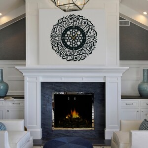 May include: A black and gold clock with Arabic calligraphy around the face, hanging on a white wall above a fireplace. The clock is in a living room setting with a white fireplace and a blue vase on a white cabinet.