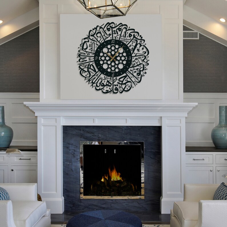 May include: A black and gold clock with Arabic calligraphy around the face, hanging on a white wall above a fireplace. The clock is in a living room setting with a white fireplace and a blue vase on a white cabinet.