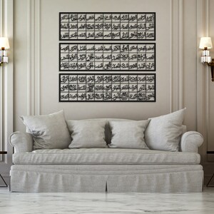 May include: Three black metal framed wall art pieces with Arabic calligraphy in a modern living room setting with a gray couch and two lamps on either side of the art.