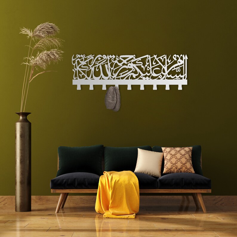 May include: A white metal wall decor featuring Arabic calligraphy and a decorative design. The decor is hanging on a green wall above a black sofa with a yellow throw and cushions.