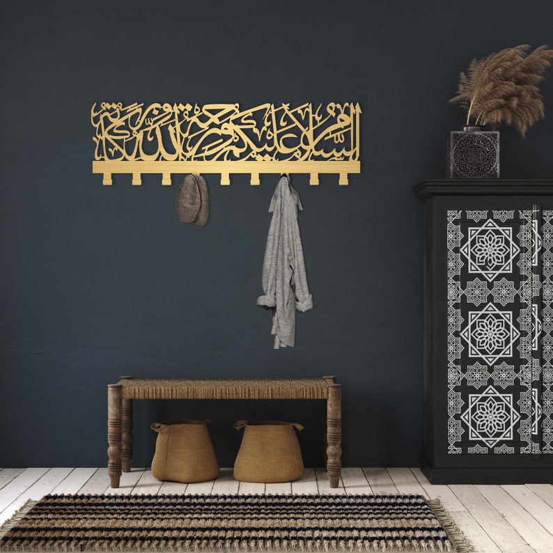 May include: A gold metal wall-mounted coat rack with Arabic calligraphy. The rack has multiple hooks and is mounted on a dark blue wall. A grey scarf and a brown hat are hanging on the hooks.