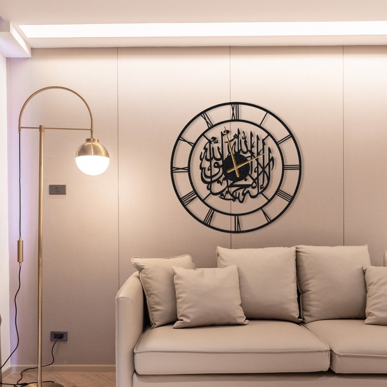 K&ouml;nnte beinhalten: Eine schwarze Metall-Wanduhr mit arabischer Kalligraphie und r&ouml;mischen Ziffern. Die Uhr ist rund und h&auml;ngt an einer beigen Wand. Eine goldene Stehlampe mit kugelf&ouml;rmigem Licht und ein beiges Sofa befinden sich im Vordergrund.